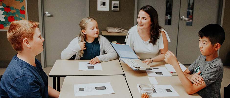 Female teacher smiles while opening a book among students sitting at their desk