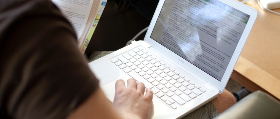 male typing on a computer while holding a textbook