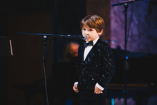 Young child dressed in a shiny tuxedo speaks into a microphone